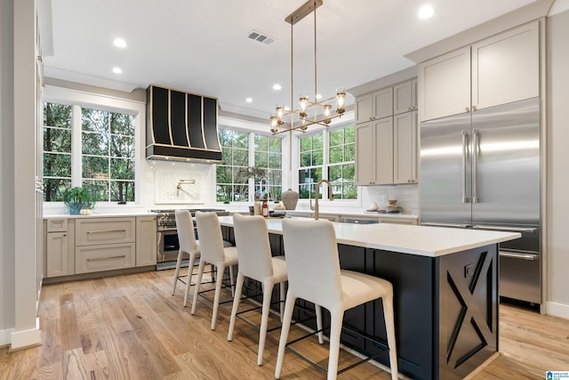 kitchen with custom exhaust hood, light hardwood / wood-style flooring, gray cabinets, an island with sink, and premium appliances