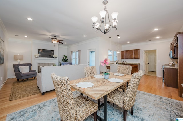 dining space with ceiling fan with notable chandelier, sink, crown molding, light wood-type flooring, and a tiled fireplace