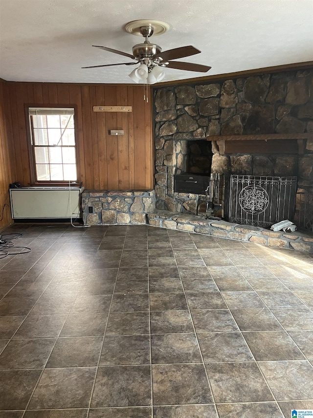 unfurnished living room with a textured ceiling, a stone fireplace, ceiling fan, and wood walls