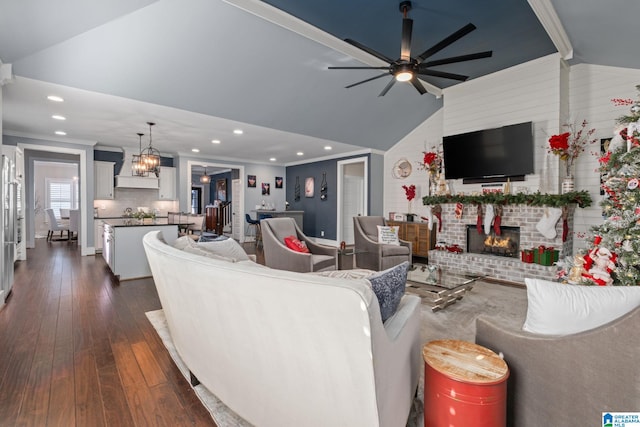 living room with dark wood-type flooring, a brick fireplace, lofted ceiling, ceiling fan with notable chandelier, and ornamental molding