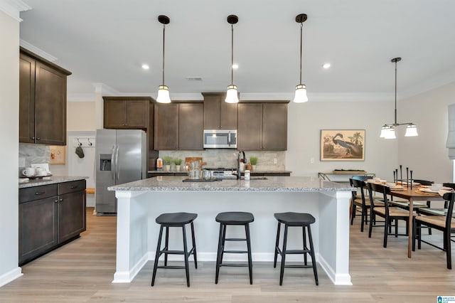 kitchen with stainless steel appliances, light stone counters, light hardwood / wood-style flooring, decorative light fixtures, and a kitchen island with sink