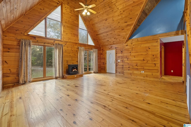 unfurnished living room featuring a wood stove, high vaulted ceiling, wooden walls, ceiling fan, and light hardwood / wood-style floors