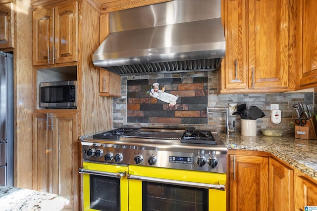 kitchen featuring wall chimney exhaust hood, light stone countertops, backsplash, and appliances with stainless steel finishes