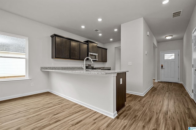 kitchen with dark brown cabinetry, light stone countertops, kitchen peninsula, appliances with stainless steel finishes, and light wood-type flooring