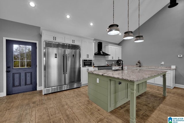 kitchen featuring wall chimney range hood, stainless steel built in refrigerator, a breakfast bar area, a kitchen island with sink, and white cabinets