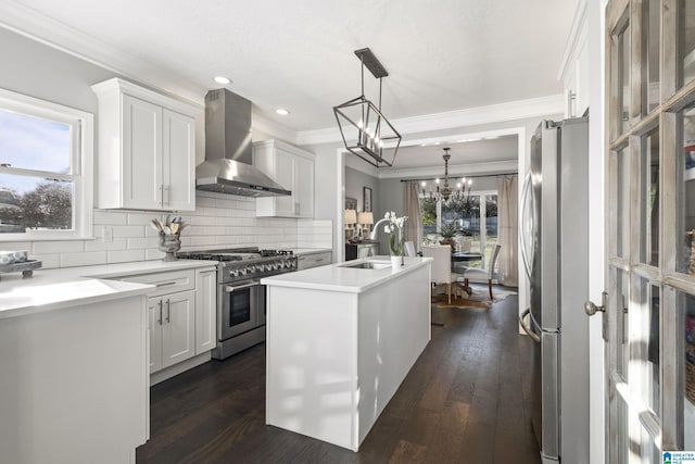 kitchen featuring wall chimney exhaust hood, stainless steel appliances, pendant lighting, a center island with sink, and white cabinetry