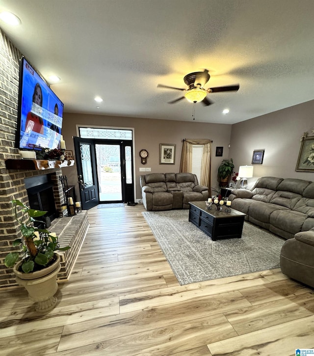 living room featuring ceiling fan, light wood-type flooring, a textured ceiling, and a brick fireplace