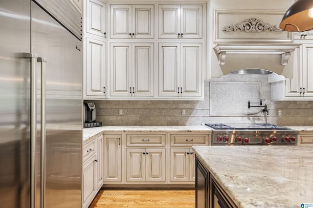 kitchen featuring high quality appliances, light stone countertops, tasteful backsplash, light hardwood / wood-style floors, and custom range hood