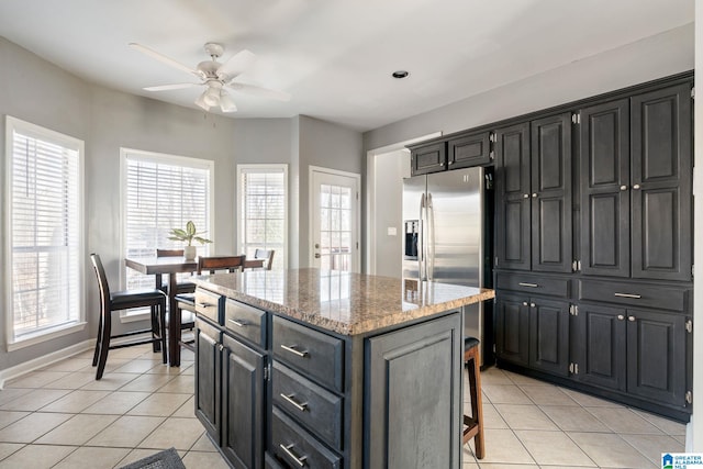 kitchen with stainless steel refrigerator with ice dispenser, ceiling fan, light stone countertops, light tile patterned floors, and a kitchen island