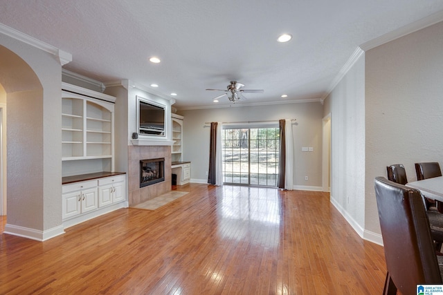 living room featuring built in shelves, ceiling fan, ornamental molding, a tiled fireplace, and light wood-type flooring