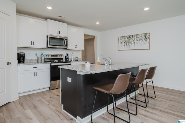 kitchen with light stone countertops, stainless steel appliances, a kitchen island with sink, light hardwood / wood-style flooring, and white cabinetry