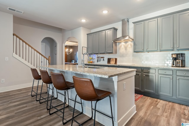 kitchen with sink, wall chimney exhaust hood, light stone countertops, an island with sink, and wood-type flooring