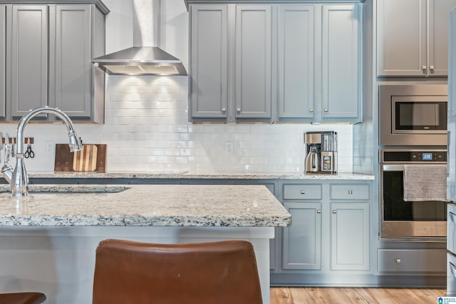 kitchen featuring wall chimney exhaust hood, light stone countertops, appliances with stainless steel finishes, and tasteful backsplash