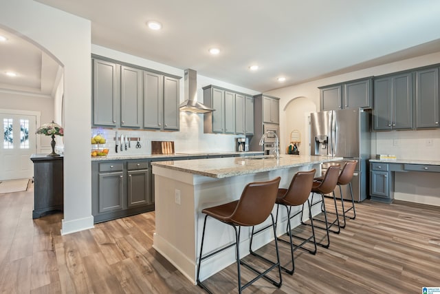 kitchen with stainless steel refrigerator with ice dispenser, gray cabinets, a kitchen island with sink, and wall chimney exhaust hood