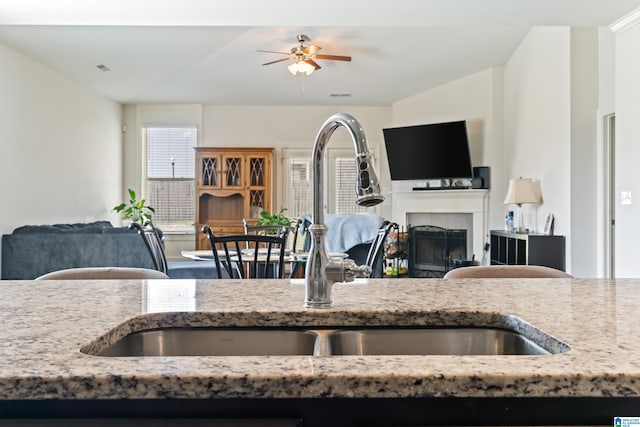 kitchen with a tile fireplace, light stone counters, ceiling fan, and sink