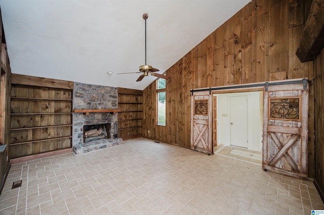 unfurnished living room with ceiling fan, wooden walls, a barn door, built in features, and a stone fireplace