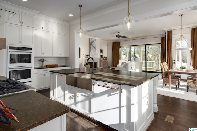 kitchen with backsplash, multiple ovens, ceiling fan, an island with sink, and white cabinetry