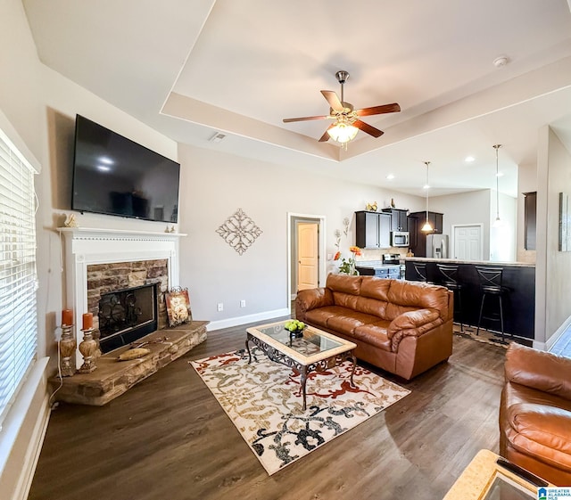 living room featuring dark hardwood / wood-style flooring, a tray ceiling, a stone fireplace, and ceiling fan