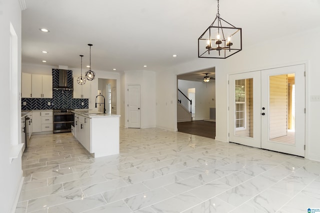 kitchen featuring french doors, a kitchen island with sink, sink, wall chimney range hood, and decorative light fixtures