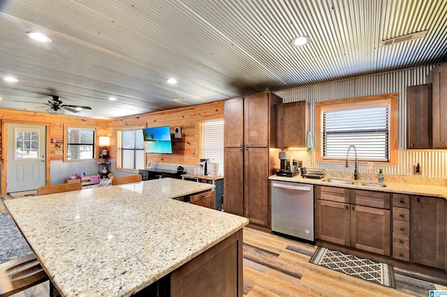 kitchen featuring light stone counters, stainless steel dishwasher, ceiling fan, wooden walls, and sink