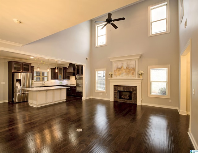 unfurnished living room featuring a towering ceiling, ceiling fan, dark wood-type flooring, sink, and a stone fireplace