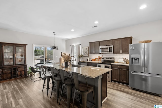 kitchen featuring sink, a kitchen island with sink, light stone counters, decorative backsplash, and appliances with stainless steel finishes