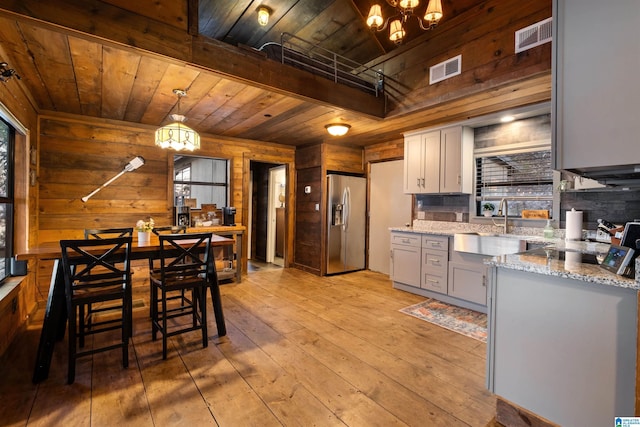 kitchen featuring sink, wooden walls, light stone counters, hanging light fixtures, and stainless steel fridge