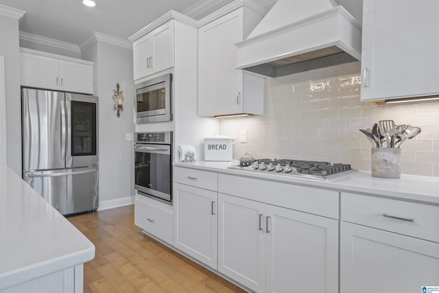 kitchen with white cabinetry, ornamental molding, custom exhaust hood, backsplash, and appliances with stainless steel finishes