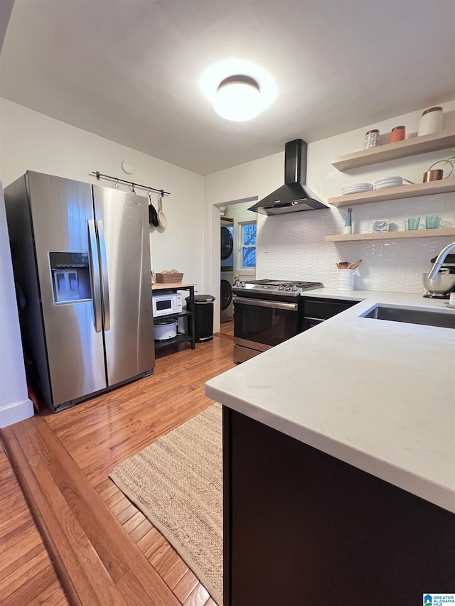 kitchen with appliances with stainless steel finishes, sink, wall chimney range hood, stacked washer and clothes dryer, and light hardwood / wood-style floors