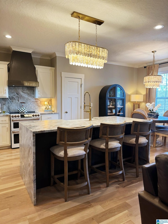 kitchen with decorative light fixtures, premium range hood, a kitchen island with sink, range with two ovens, and white cabinets