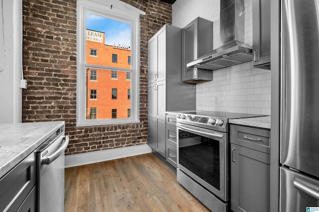 kitchen featuring brick wall, appliances with stainless steel finishes, wall chimney range hood, and gray cabinets