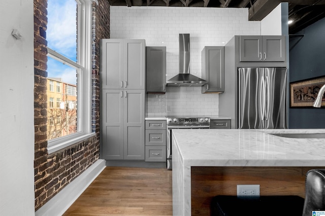 kitchen with light stone countertops, gray cabinetry, stainless steel appliances, wall chimney exhaust hood, and brick wall