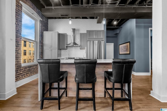 kitchen with gray cabinets, a kitchen breakfast bar, wall chimney exhaust hood, stainless steel fridge, and brick wall