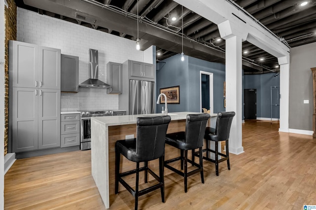 kitchen with wall chimney range hood, a kitchen island with sink, light stone countertops, appliances with stainless steel finishes, and gray cabinetry