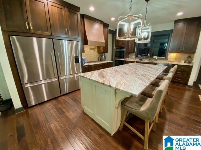 kitchen with stainless steel appliances, premium range hood, light stone counters, a kitchen island, and dark brown cabinetry