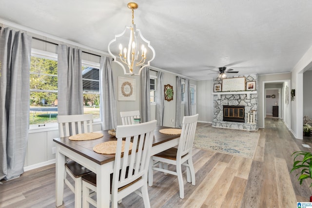 dining area with light wood-type flooring, a stone fireplace, a wealth of natural light, and ceiling fan with notable chandelier