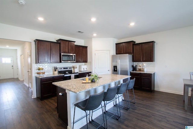 kitchen with appliances with stainless steel finishes, dark brown cabinets, an island with sink, a kitchen bar, and dark hardwood / wood-style flooring