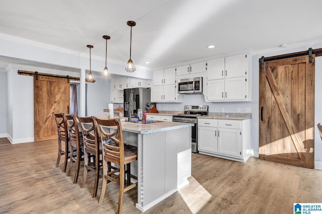 kitchen with a barn door, appliances with stainless steel finishes, a kitchen island with sink, decorative light fixtures, and white cabinets