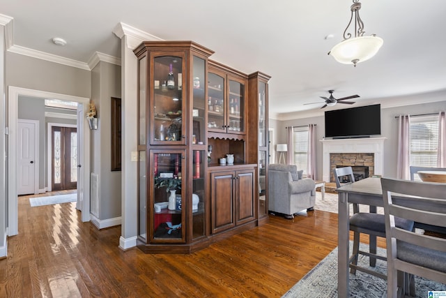 dining space with ceiling fan, dark wood-type flooring, ornamental molding, and a stone fireplace