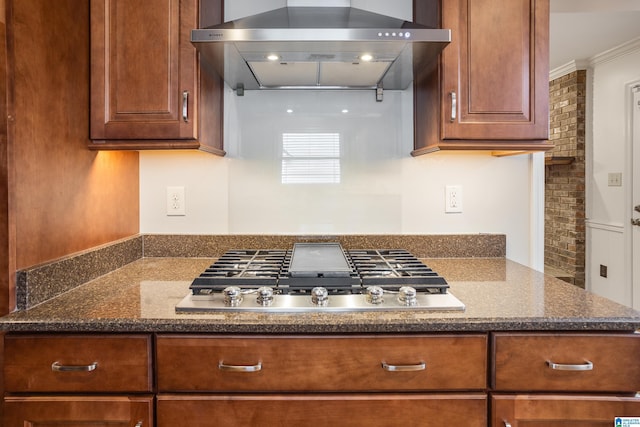 kitchen with stainless steel gas cooktop, wall chimney exhaust hood, dark stone counters, and brown cabinets