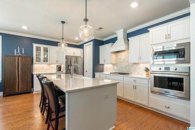 kitchen with white cabinets, stainless steel appliances, custom range hood, and a center island with sink