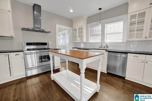 kitchen featuring appliances with stainless steel finishes, white cabinetry, wall chimney range hood, dark hardwood / wood-style flooring, and hanging light fixtures