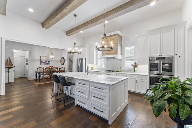 kitchen with appliances with stainless steel finishes, a kitchen island with sink, hanging light fixtures, white cabinetry, and light stone counters