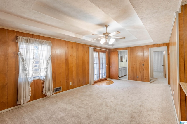 unfurnished bedroom featuring ensuite bath, light carpet, wooden walls, ceiling fan, and a tray ceiling