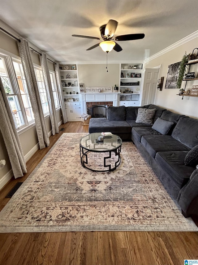 living room with ceiling fan, a fireplace, wood-type flooring, crown molding, and built in shelves