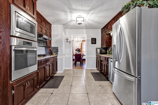 kitchen with appliances with stainless steel finishes, decorative backsplash, light tile patterned flooring, a chandelier, and dark brown cabinetry