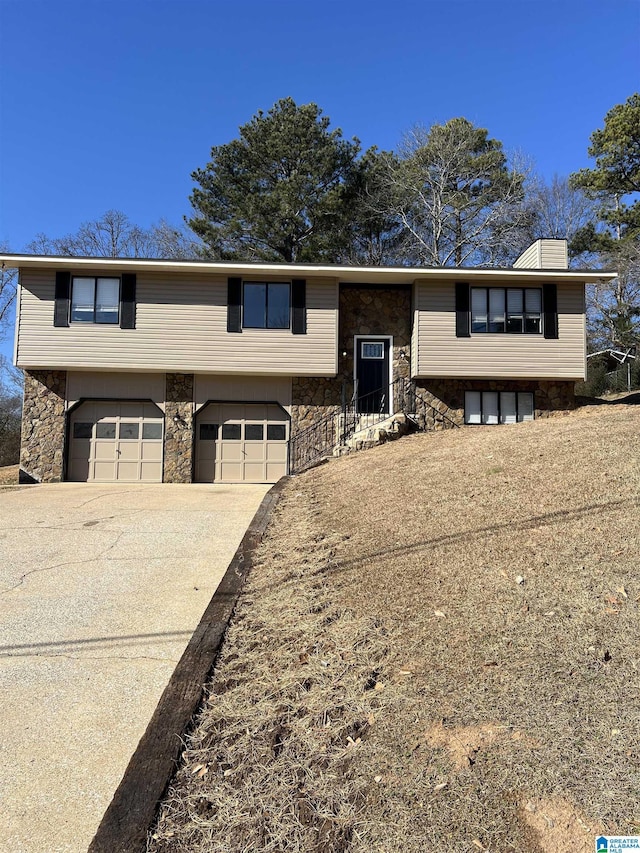 raised ranch with stone siding, driveway, a chimney, and an attached garage