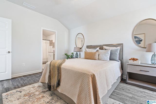 bedroom featuring wood finished floors, visible vents, baseboards, vaulted ceiling, and ensuite bath