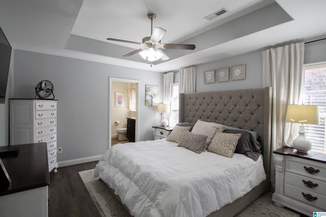 bedroom with connected bathroom, a tray ceiling, dark wood-type flooring, and ceiling fan