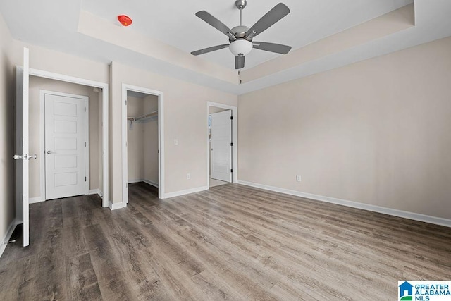 unfurnished bedroom featuring wood-type flooring, a spacious closet, a tray ceiling, a closet, and ceiling fan